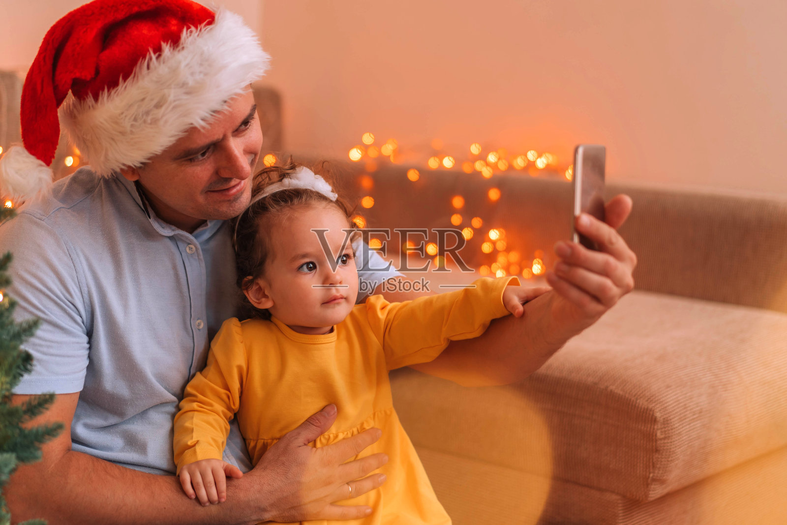 戴着红色圣诞帽的爸爸在用智能手机和可爱的小女儿自拍。照片摄影图片