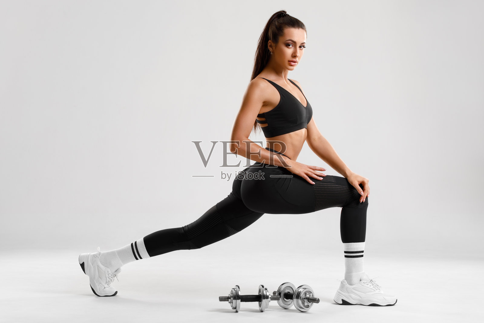 健身妇女做弓步运动腿部肌肉锻炼训练。活泼的女孩在灰色的背景下，做着前步单腿弓步运动照片摄影图片