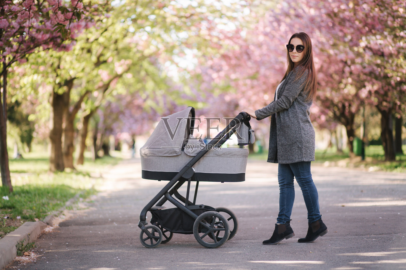 快乐的妈妈带着婴儿车里的小女孩散步。背景是粉红色的樱花树照片摄影图片