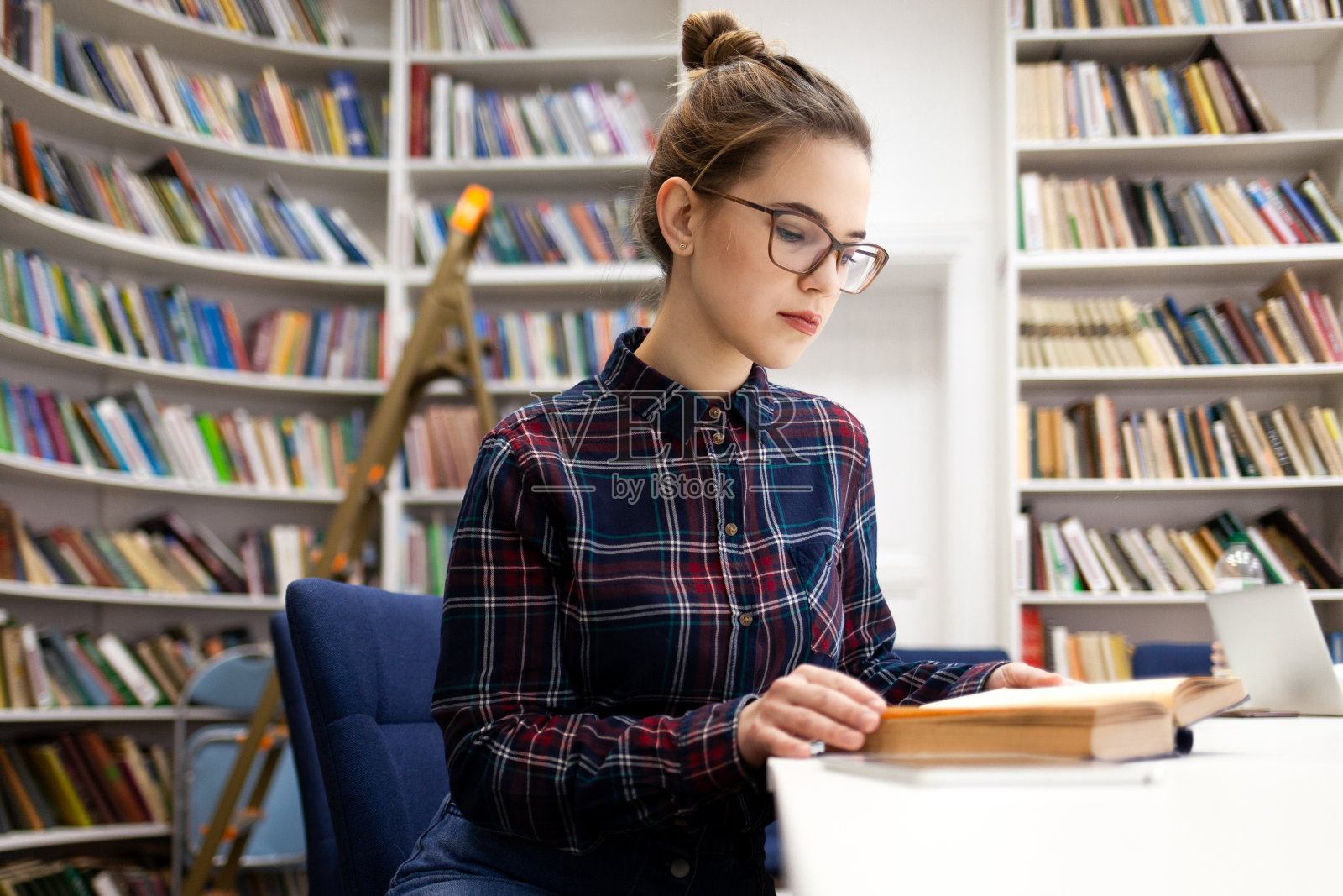 一个穿着格子衬衫的女学生坐在图书馆里翻看一本书。年轻女子来到图书馆准备大学考试。戴眼镜的女孩看书照片摄影图片