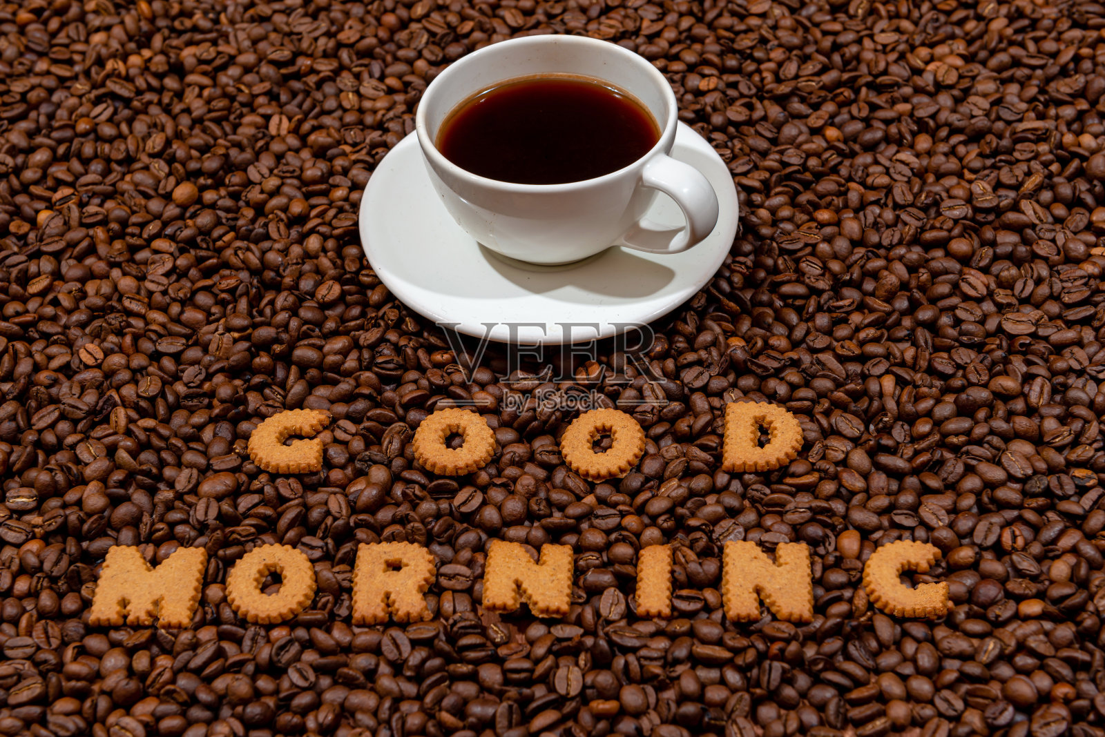 白色的咖啡杯和用饼干做成的字母写的早上好，背景是深色的咖啡豆照片摄影图片