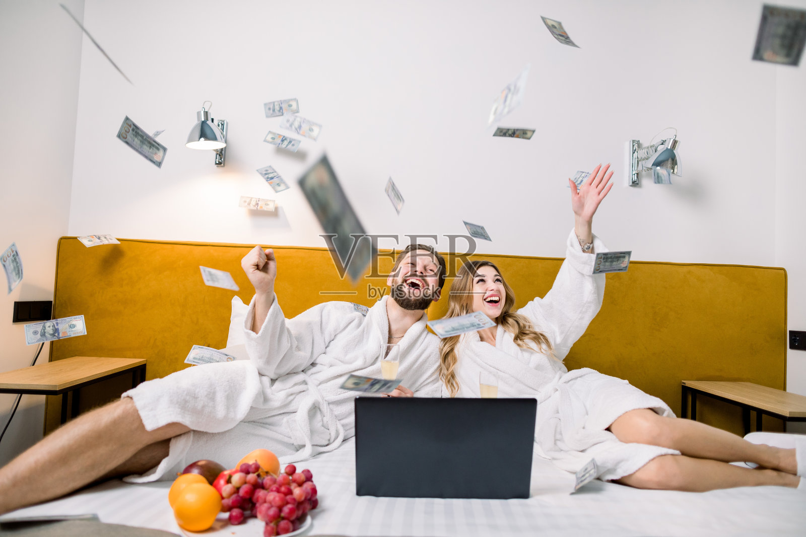 想象一对可爱的新婚夫妇坐在酒店房间的床上，一边用笔记本电脑一边吃水果，一边扔钱照片摄影图片