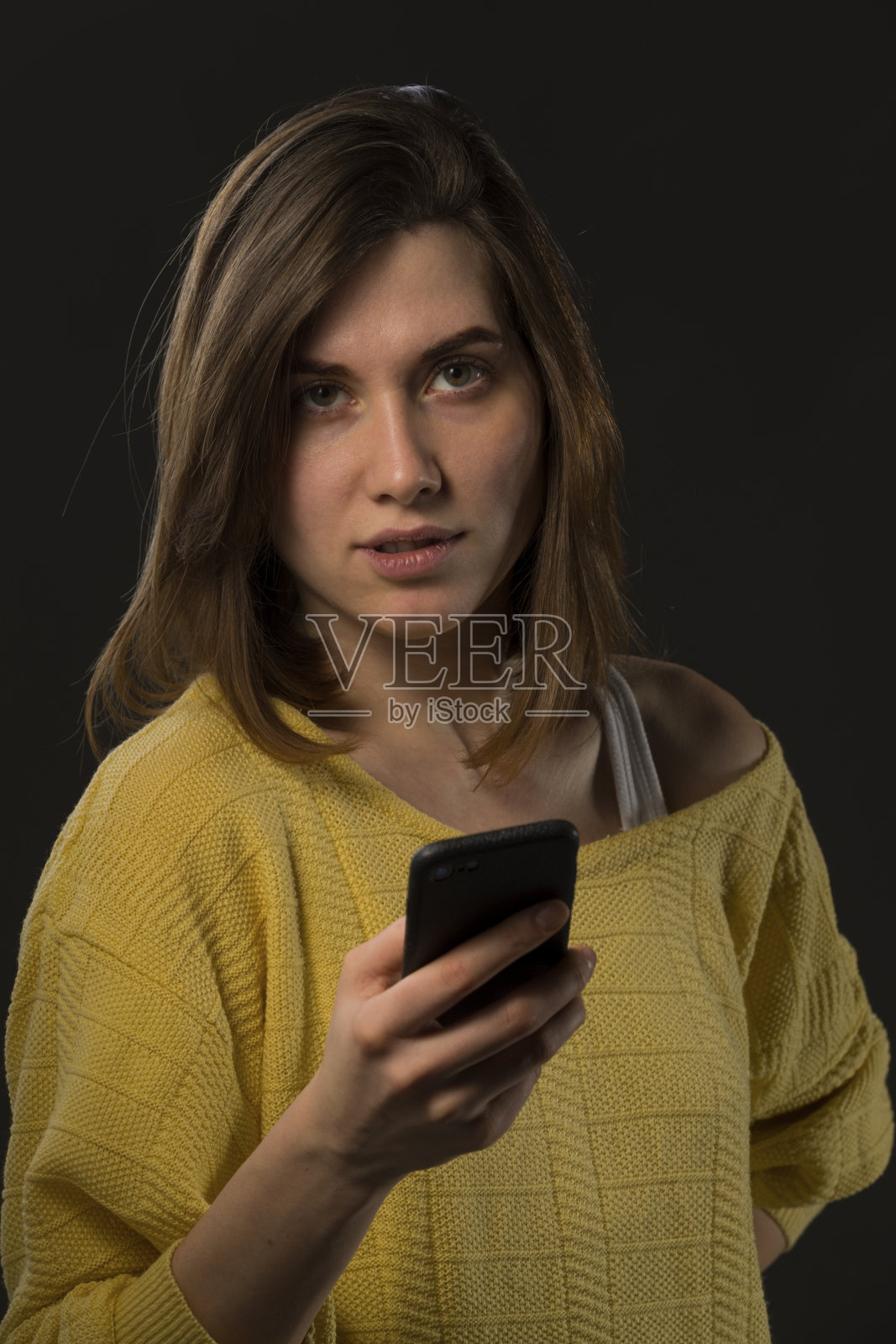 年轻情绪化的女孩穿着黄色毛衣拿着智能手机，背景是黑色照片摄影图片