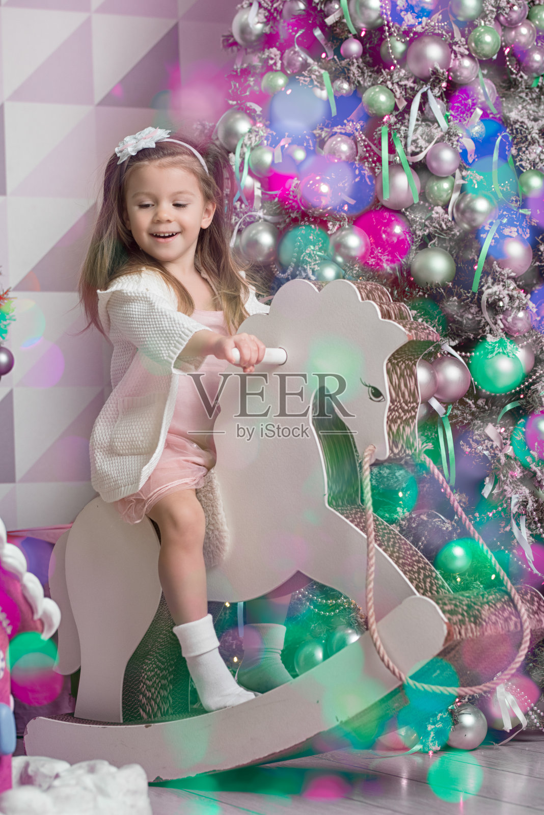 可爱的孩子(一个三岁的女孩)坐在木马上，靠着一棵新的圣诞树。散景节日灯。节日贺卡。照片摄影图片