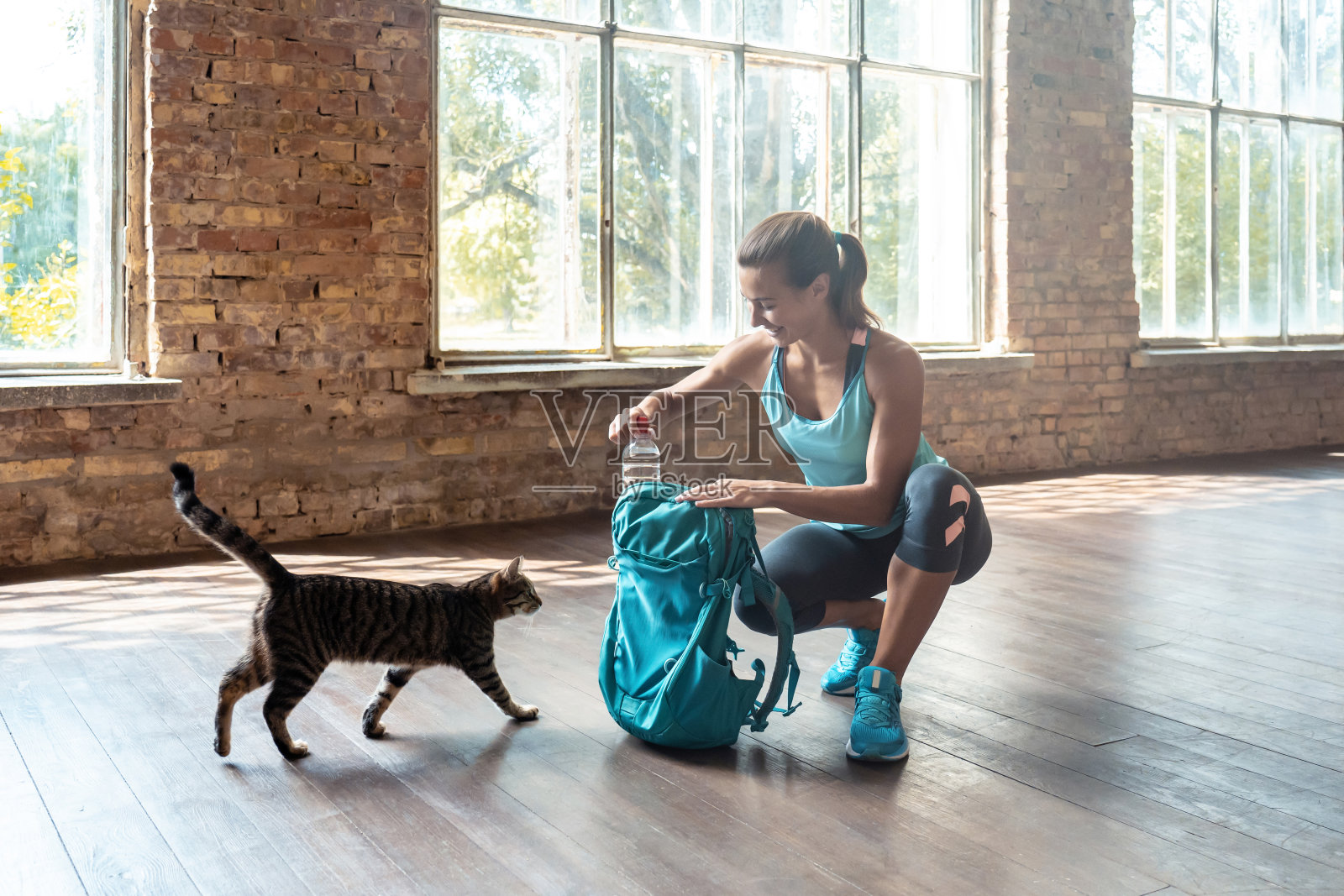 快乐健康的运动年轻女性健身教练穿着运动服，抱着水瓶打开背包在现代健身房的木地板上玩可爱有趣的猫，锻炼活跃的生活方式和动物照片摄影图片