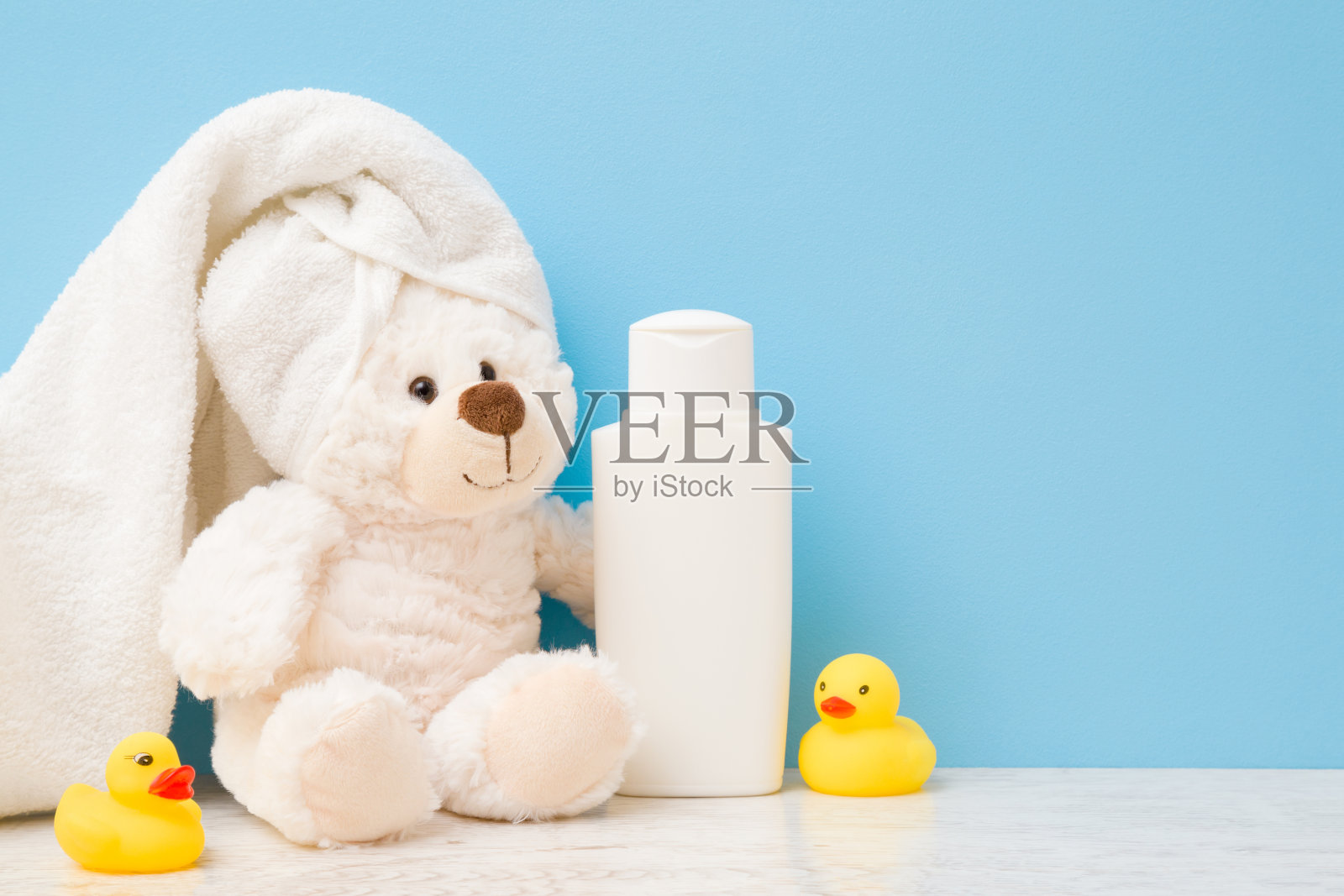 微笑着，可爱的白色泰迪熊坐着。毛巾在头上。架子上有黄色的橡皮鸭和香波瓶子。孩子洗澡的概念。浅蓝色墙壁上的文字空白位置。柔和的颜色。特写镜头。照片摄影图片