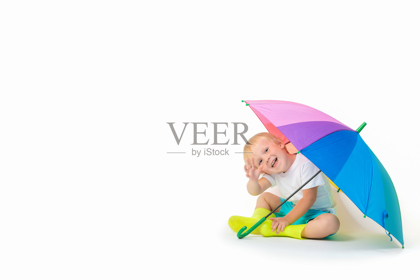 一个金发的白种人小孩坐在彩虹色的伞下，穿着胶靴，在白色的背景上挥舞着他的手。照片摄影图片