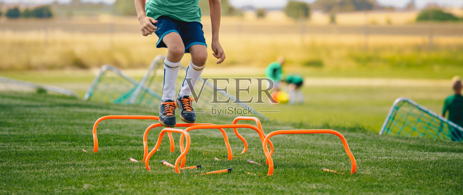 低部分肖像的不认识的男孩跳过跨栏在足球场。用足球器材训练的年轻运动员。足球速度耐力训练照片摄影图片