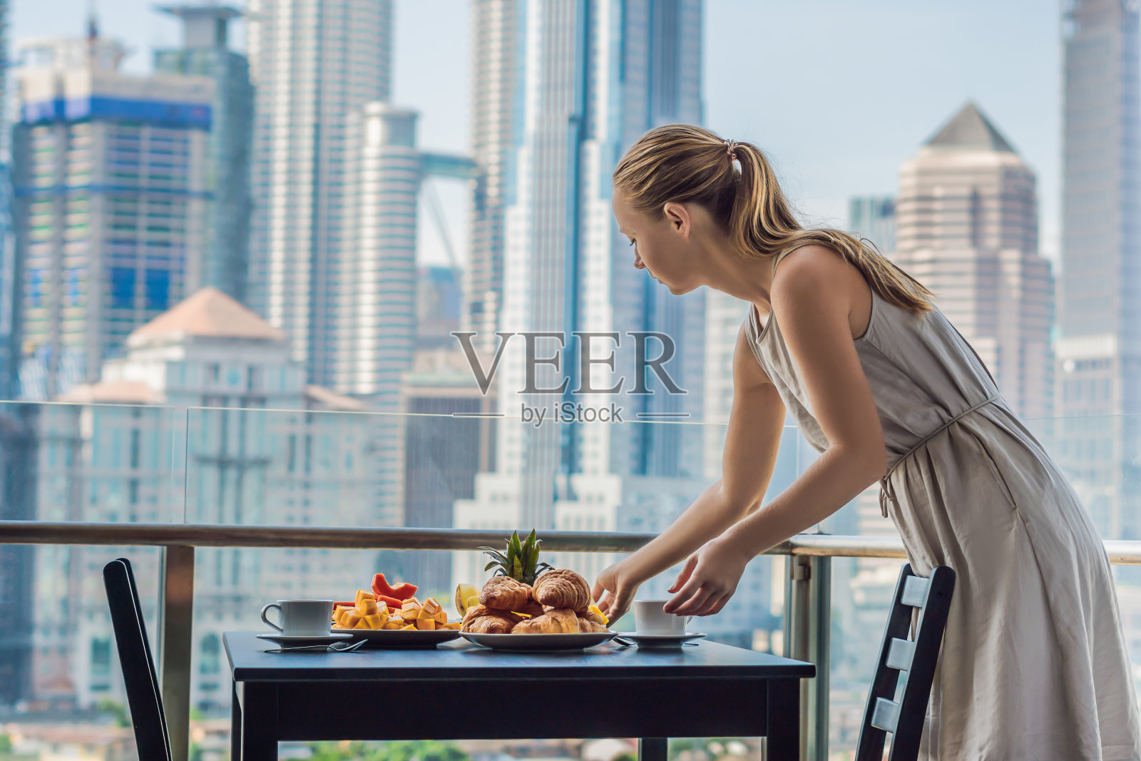 年轻女子躺在桌子上。阳台上的早餐桌上放着咖啡、水果和牛角面包，背景是大城市照片摄影图片