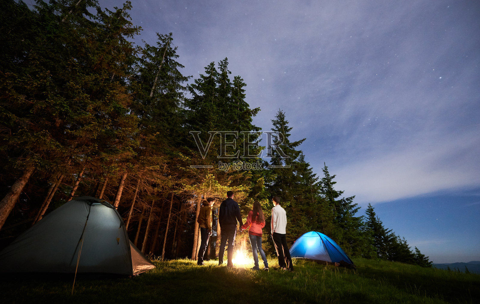 一群游客站在燃烧的帐篷旁，背景是满天星斗下的松树林照片摄影图片