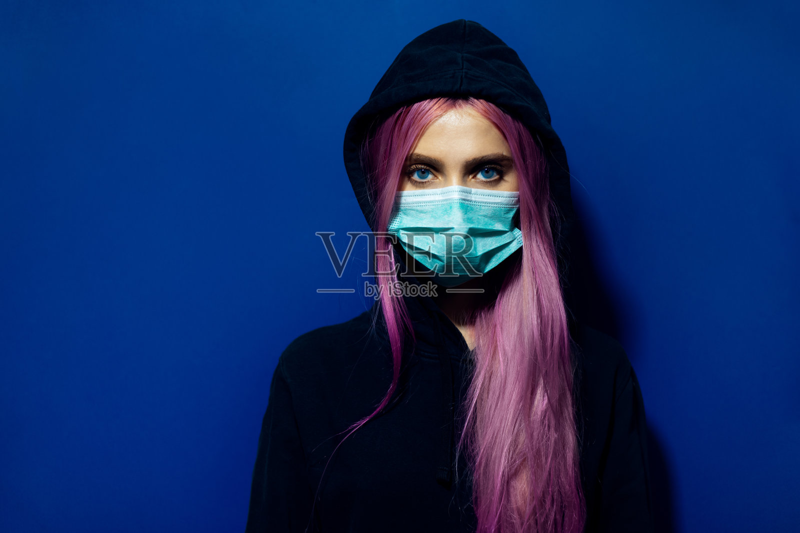 摄影棚里的年轻女孩的肖像，粉红色的头发和蓝色的眼睛，戴着医用流感口罩和连帽毛衣，背景是幻影的蓝色。照片摄影图片