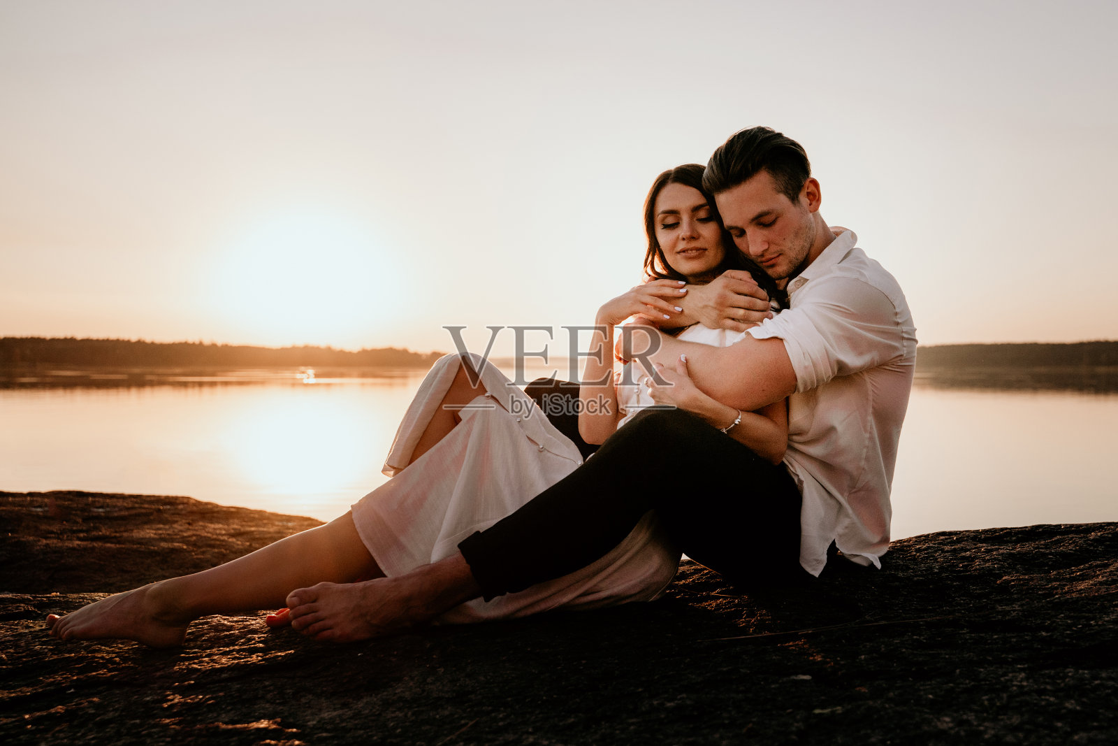 年轻夫妇拥抱和亲吻在日落海岸。浪漫的爱情故事。穿着浅色裙子的黑发女孩照片摄影图片