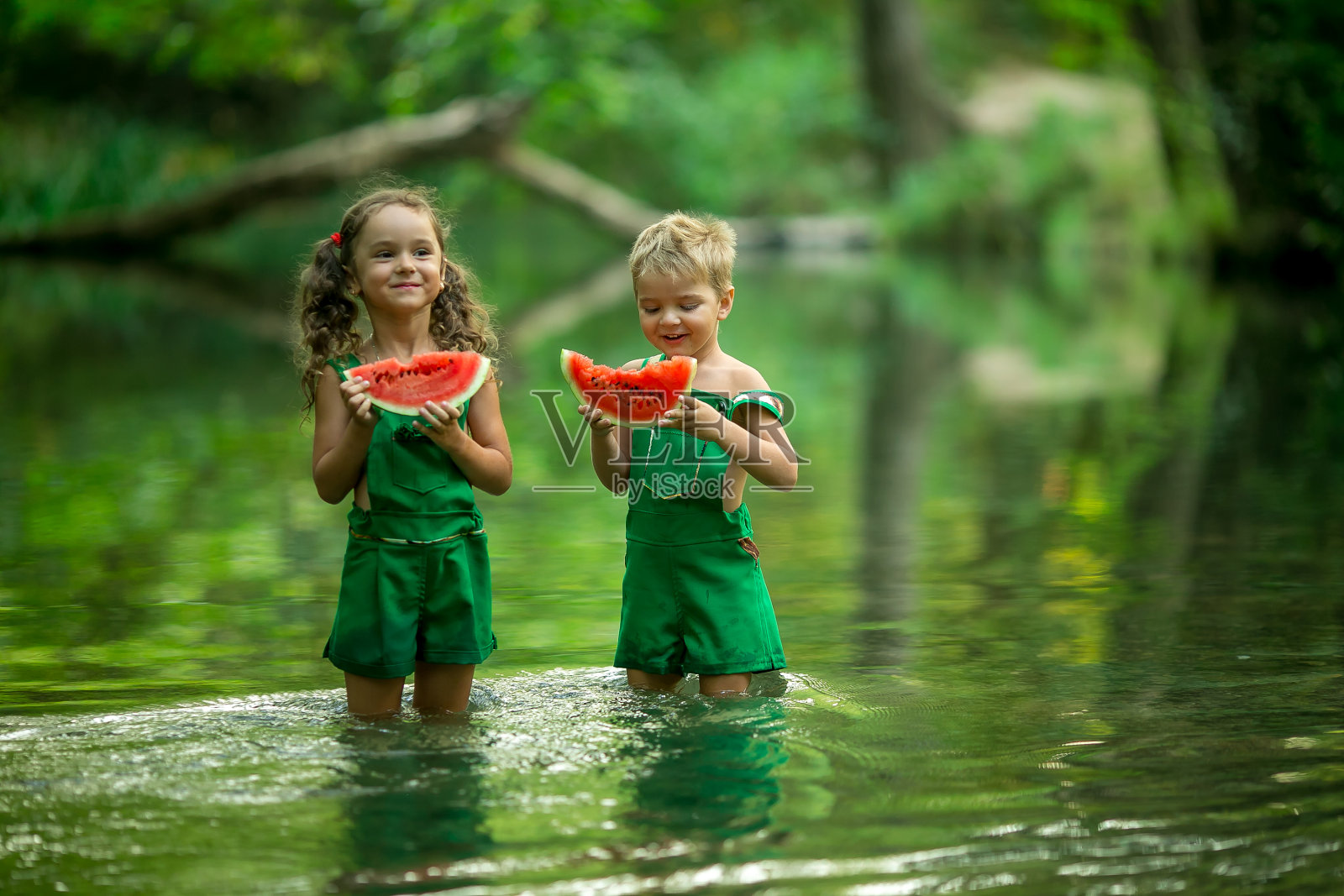 男孩和女孩站在深及膝盖的树林里的湖里吃西瓜照片摄影图片