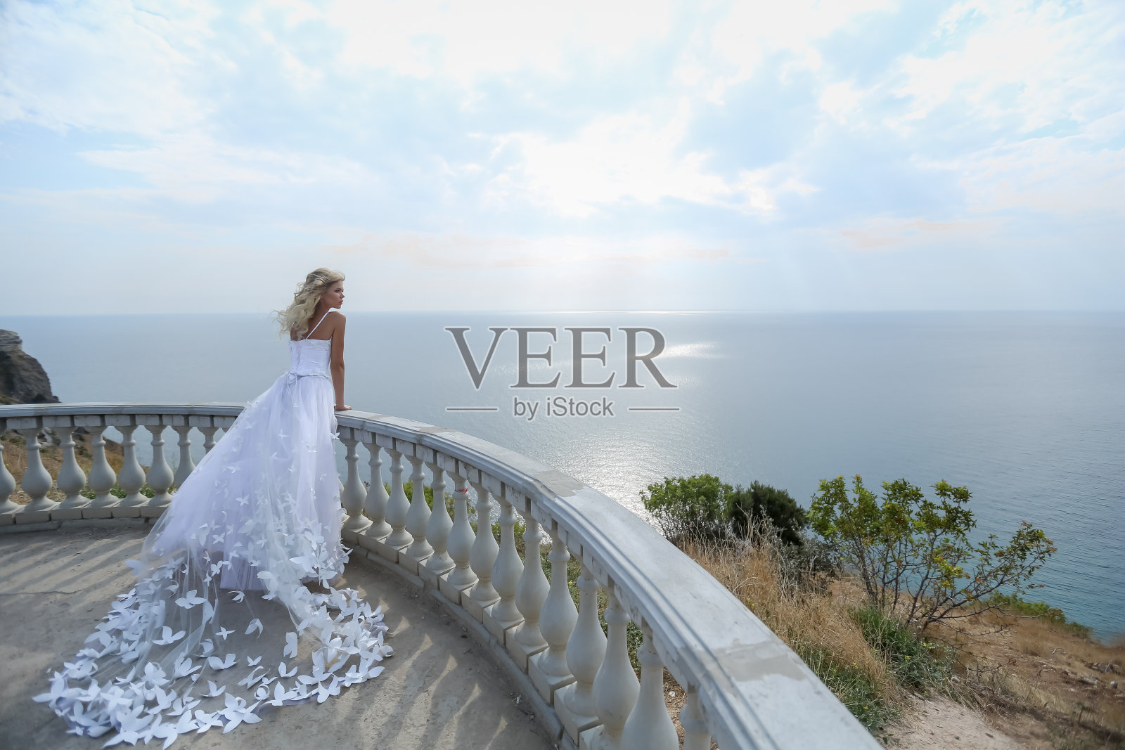 在一个阳光明媚的夏日，一位新娘穿着美丽的白色婚纱站在山顶的阳台上照片摄影图片
