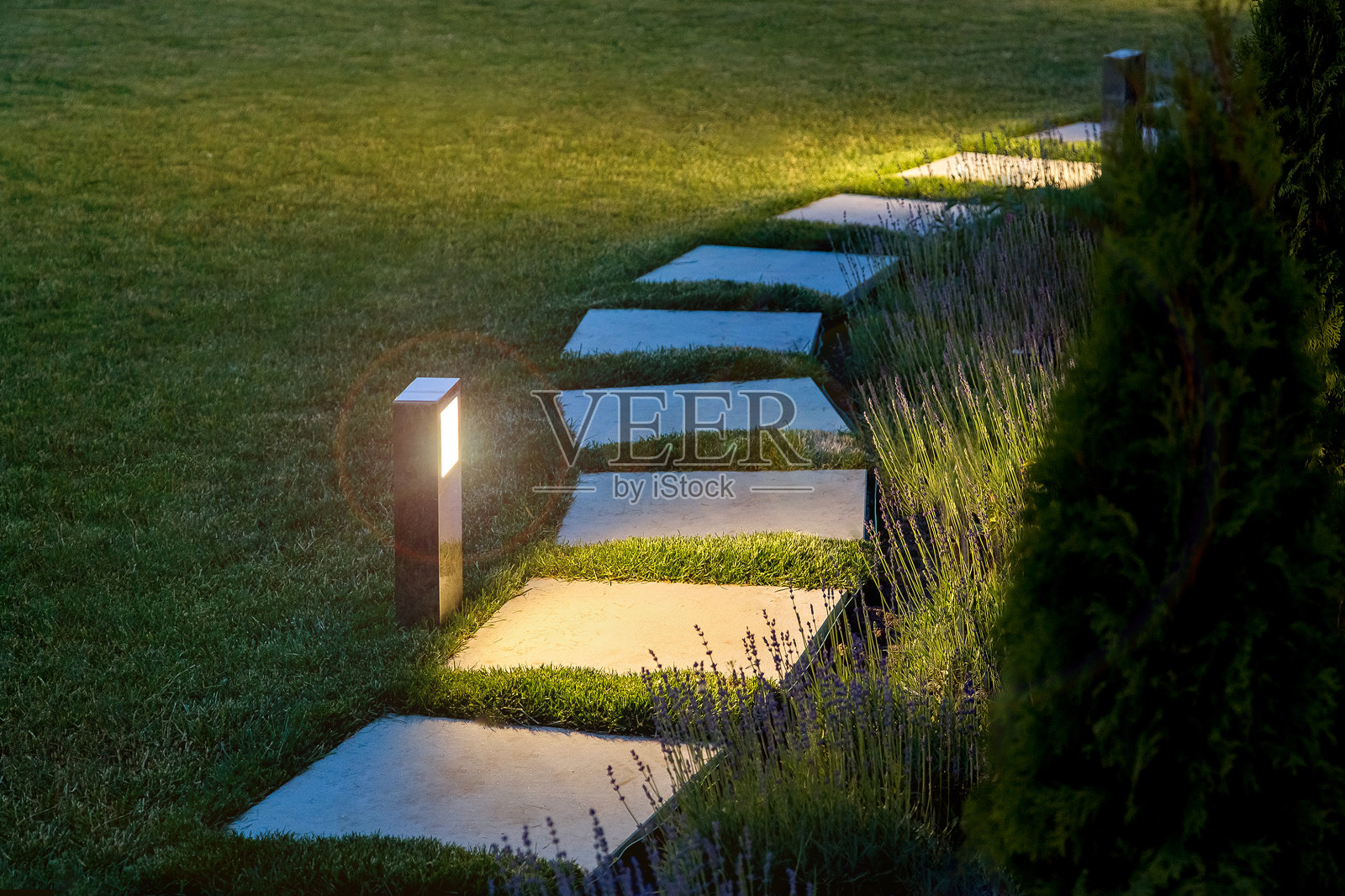 在一个有花坛和草坪的夜晚花园里，一盏温暖的灯照亮了方砖铺成的大理石小路，没有人。照片摄影图片