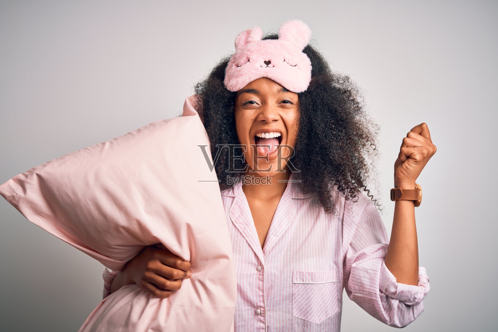 年轻的非洲裔美国妇女，留着非洲式发型，穿着睡衣，戴着睡眠面罩，抱着枕头尖叫着庆祝胜利和成功非常激动，欢呼的情绪照片摄影图片
