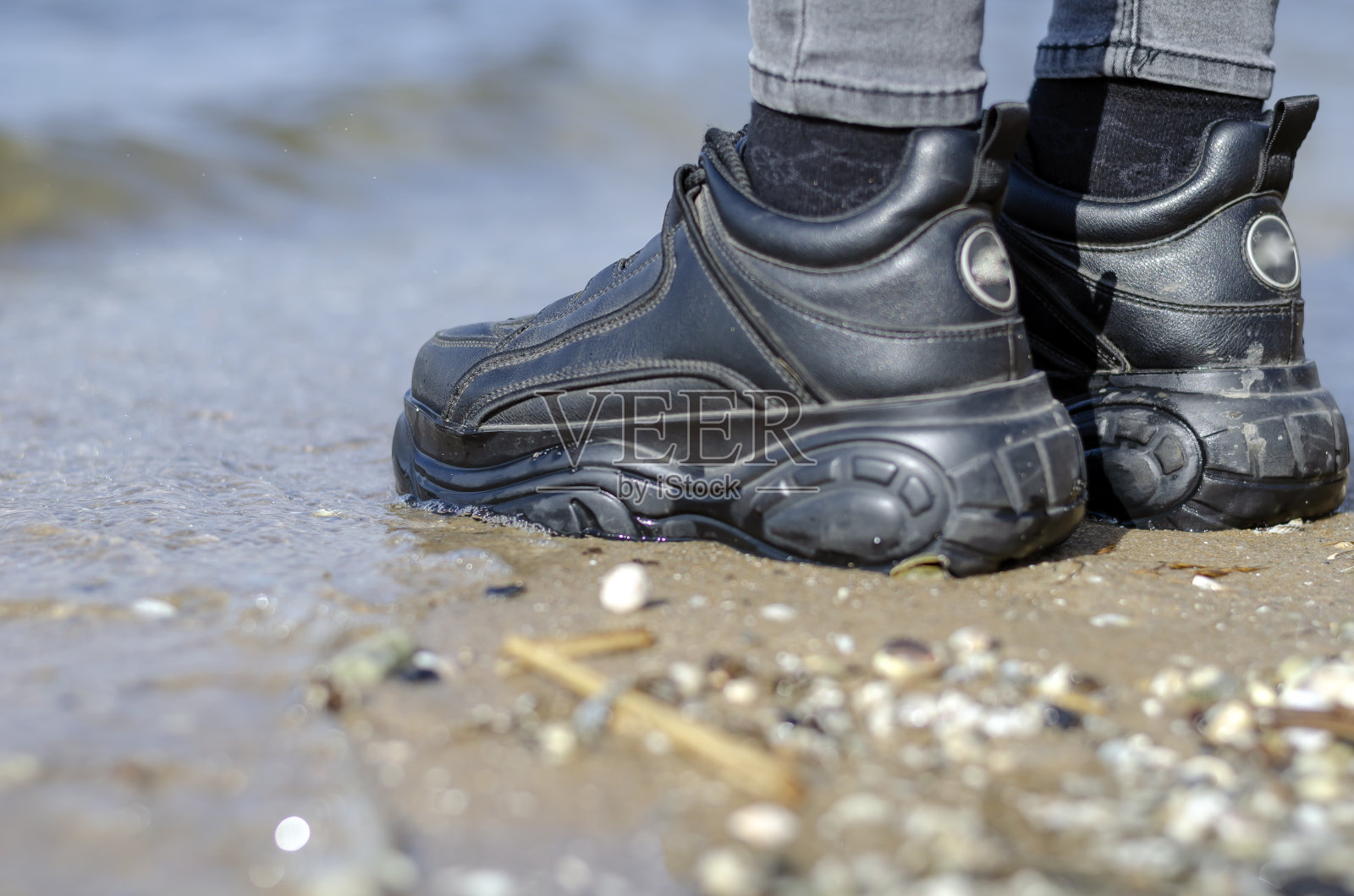 一个穿着黑色大运动鞋的女人的腿在冲浪线附近潮湿的沙滩上。照片摄影图片