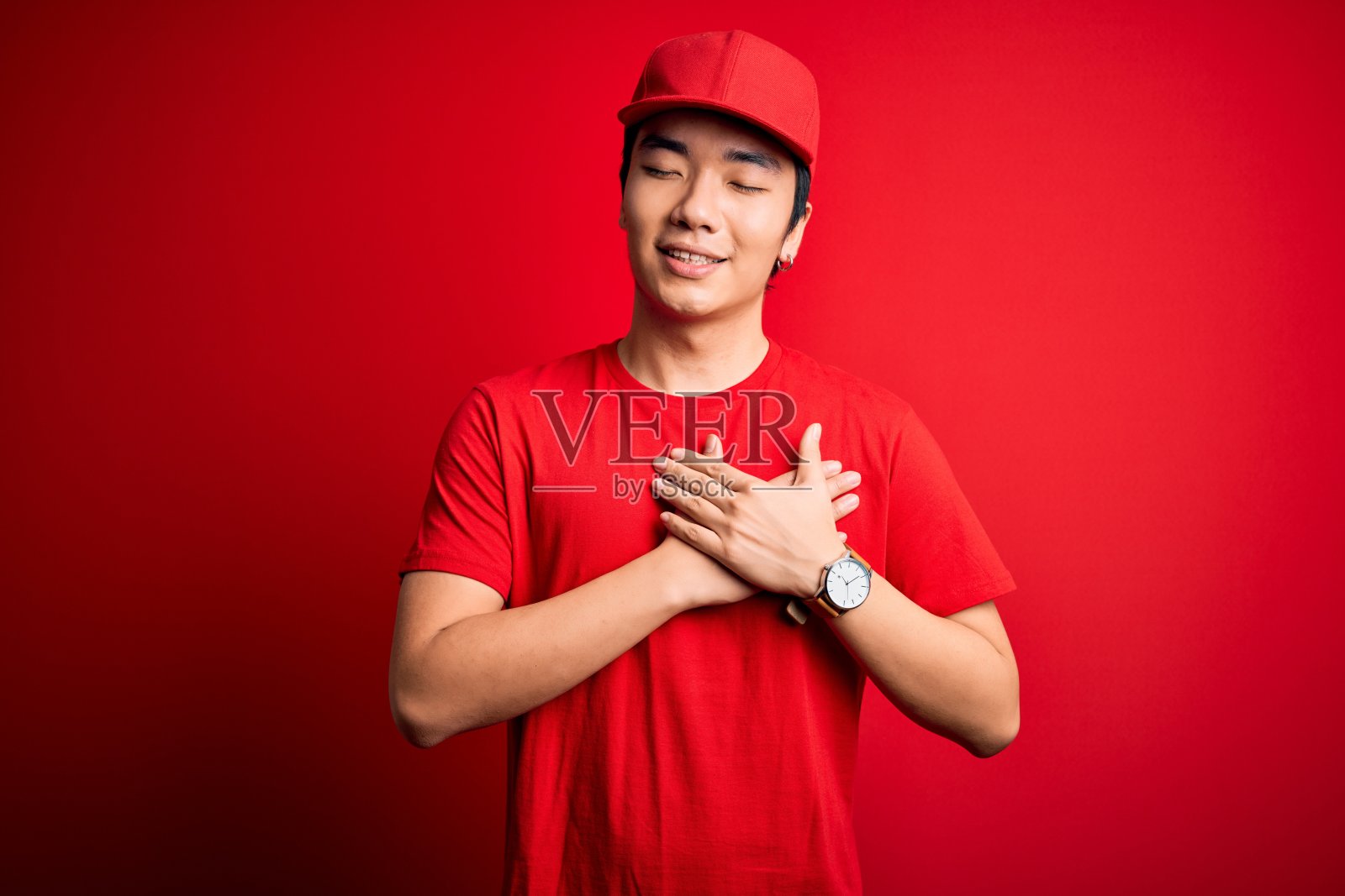 年轻英俊的中国快递员戴着帽子站在孤立的红色背景上微笑，双手放在胸前，闭上眼睛，脸上有感激的手势。健康的概念。照片摄影图片