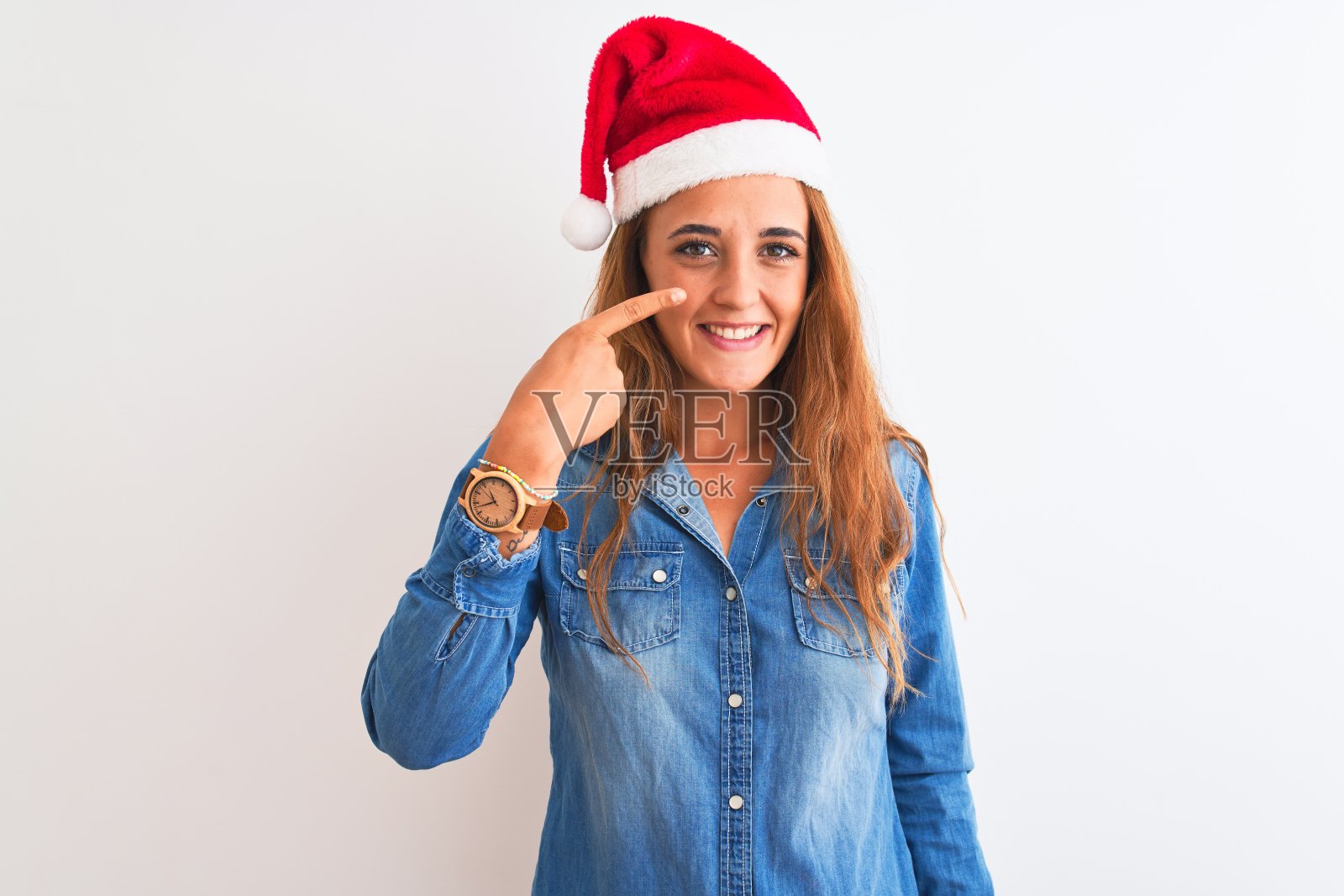 年轻漂亮的红头发女人戴着圣诞帽在孤立的背景指向与手手指的脸和鼻子，微笑愉快。美丽的概念照片摄影图片