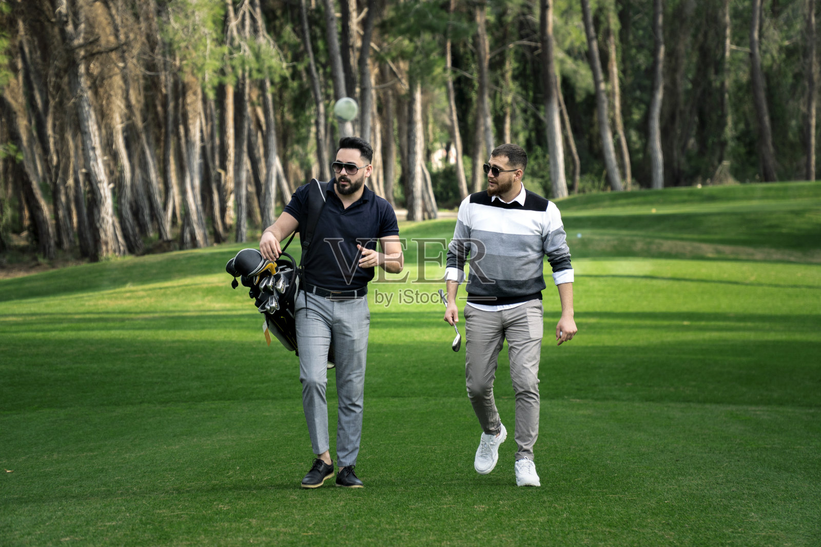 这是两名男性高尔夫球手在高尔夫球场中背着高尔夫球袋沿着球道行走的照片照片摄影图片