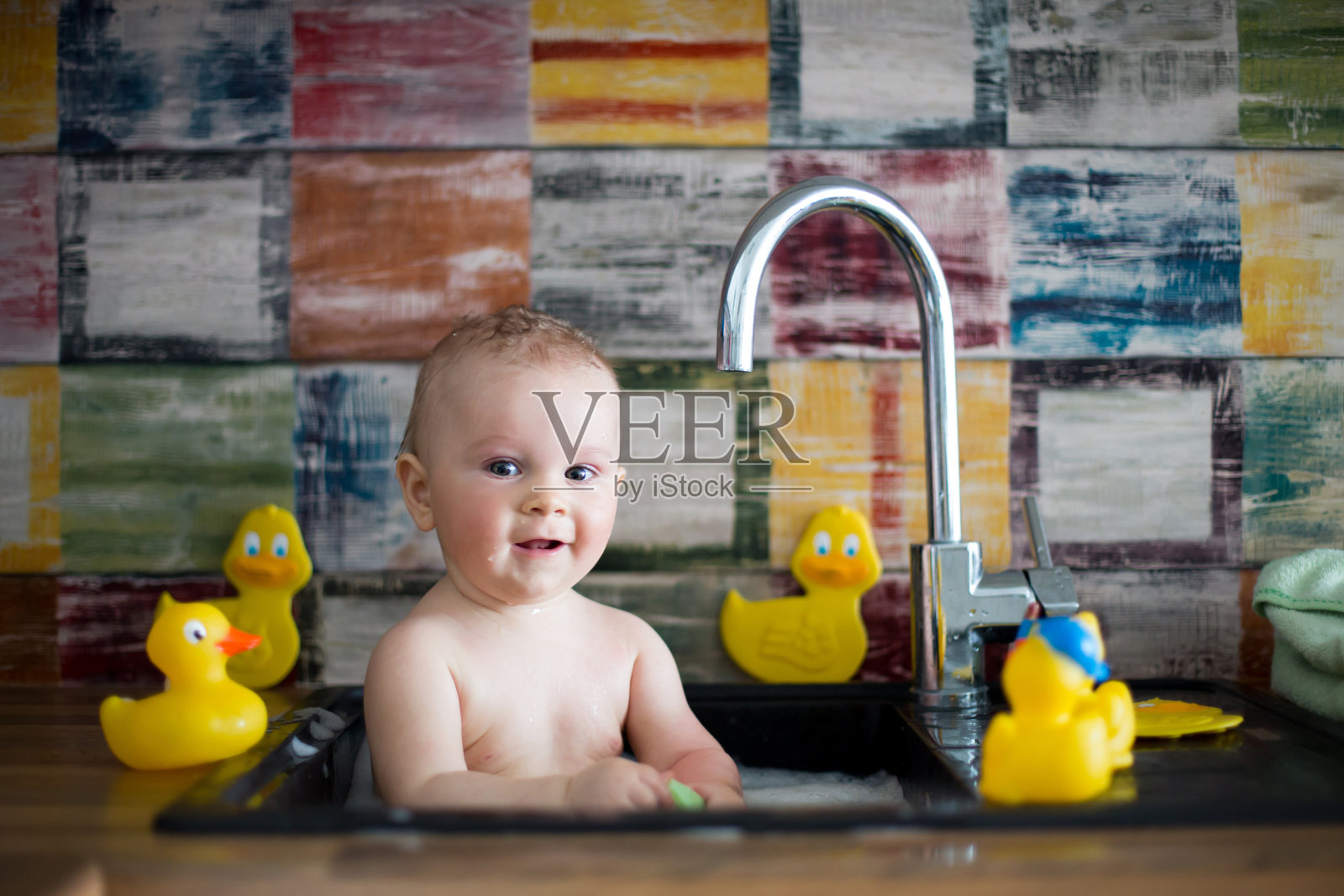 可爱的婴儿在厨房的水槽洗澡。孩子在阳光明媚的厨房里和橡皮鸭一起玩泡沫和肥皂泡，小男孩在洗澡照片摄影图片