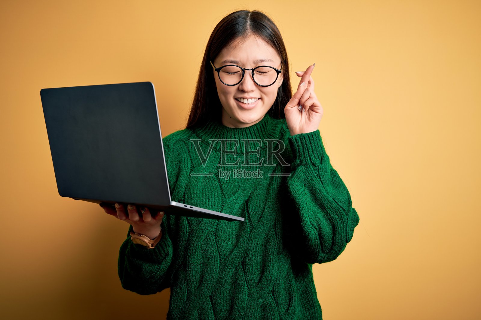 年轻的亚洲商务女性，戴着眼镜，用电脑工作，手指交叉，微笑，希望和闭上眼睛。运气和迷信的概念。照片摄影图片