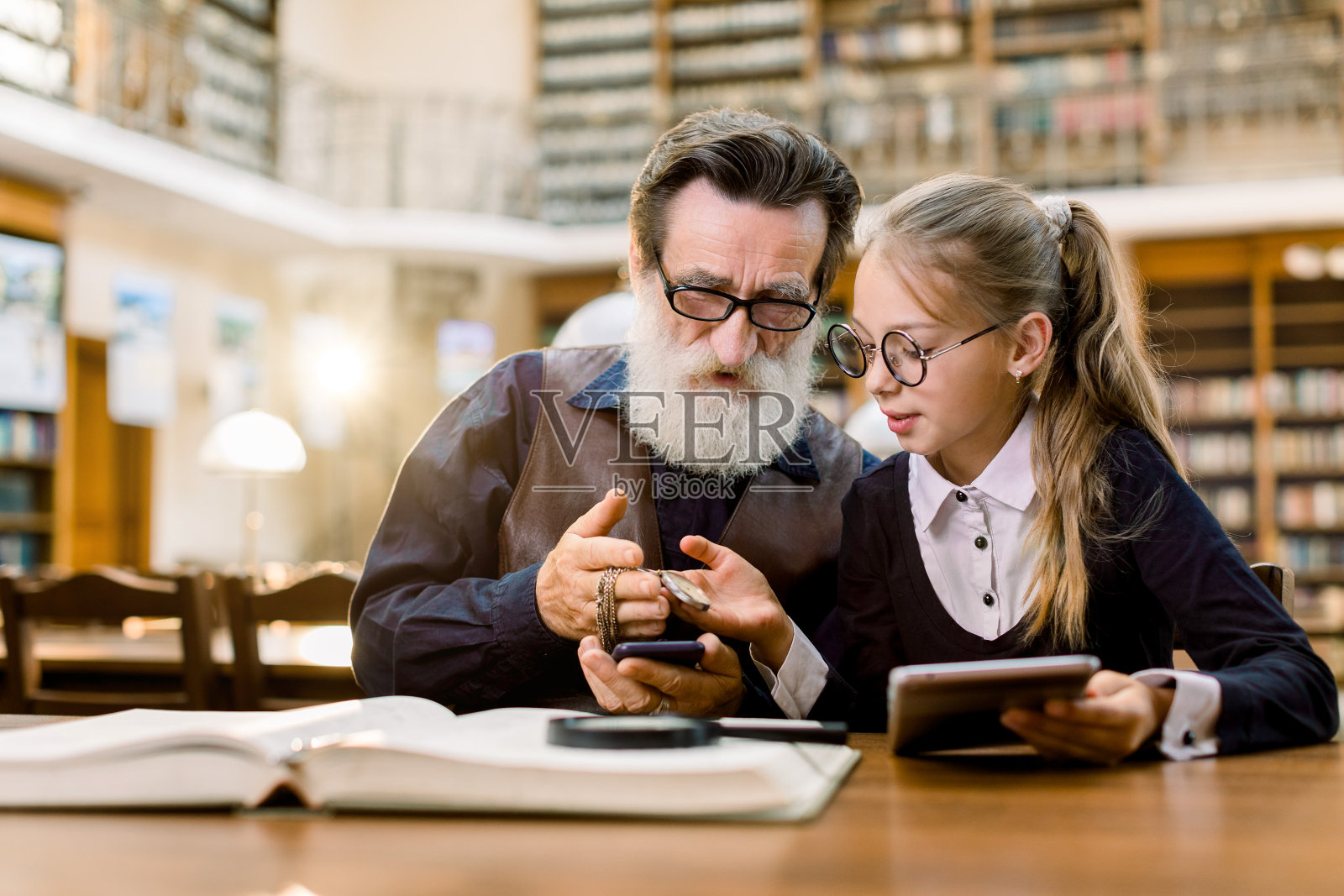 长着胡子的老帅哥和他漂亮的孙女看着一个挂在链条上的古董钟，而坐在桌子上的书，平板电脑和手机在古老的图书馆。照片摄影图片