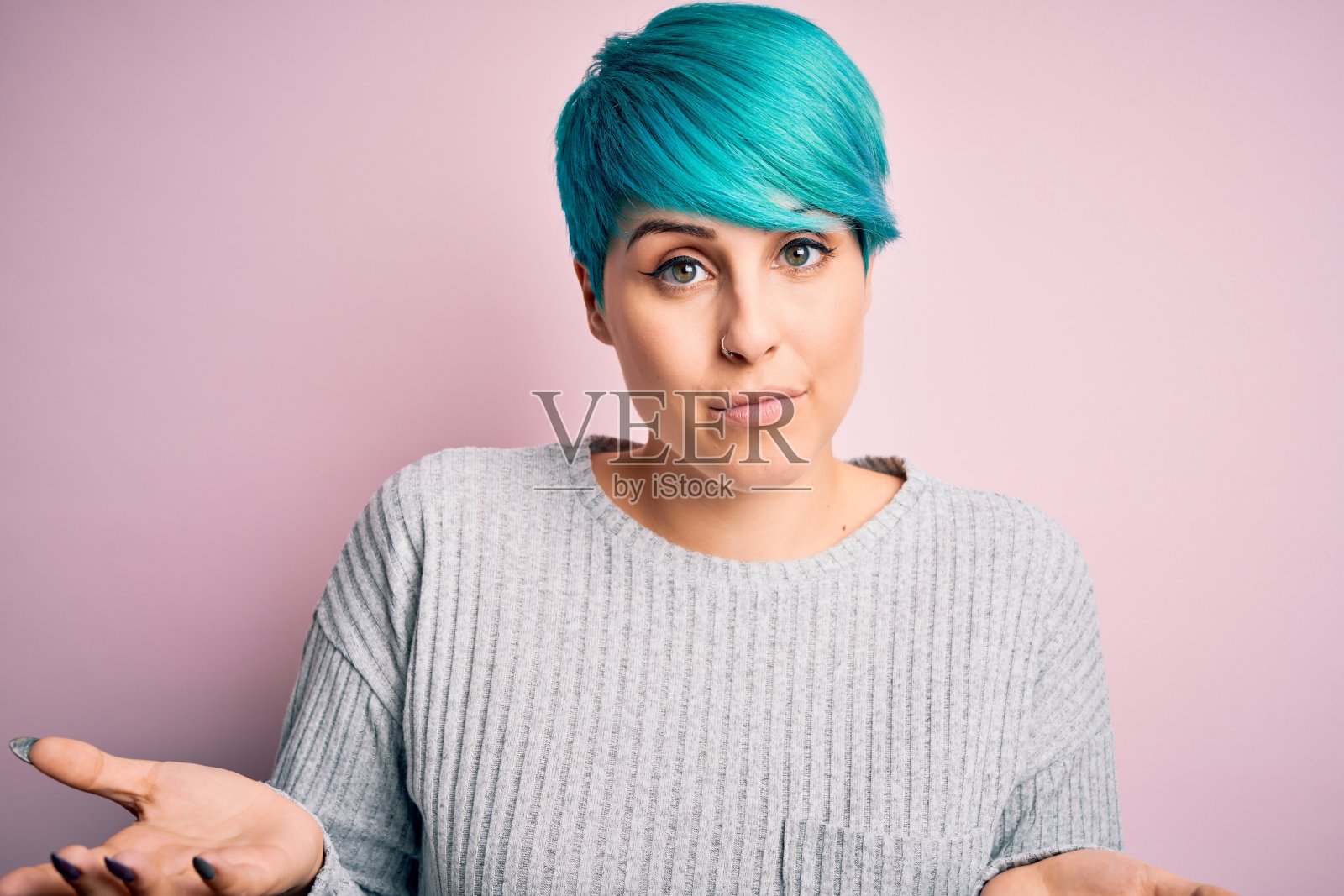 年轻美丽的女人与蓝色时尚的头发穿着休闲毛衣在粉红色的背景愚蠢和困惑的表情与胳膊和手举起。怀疑的概念。照片摄影图片