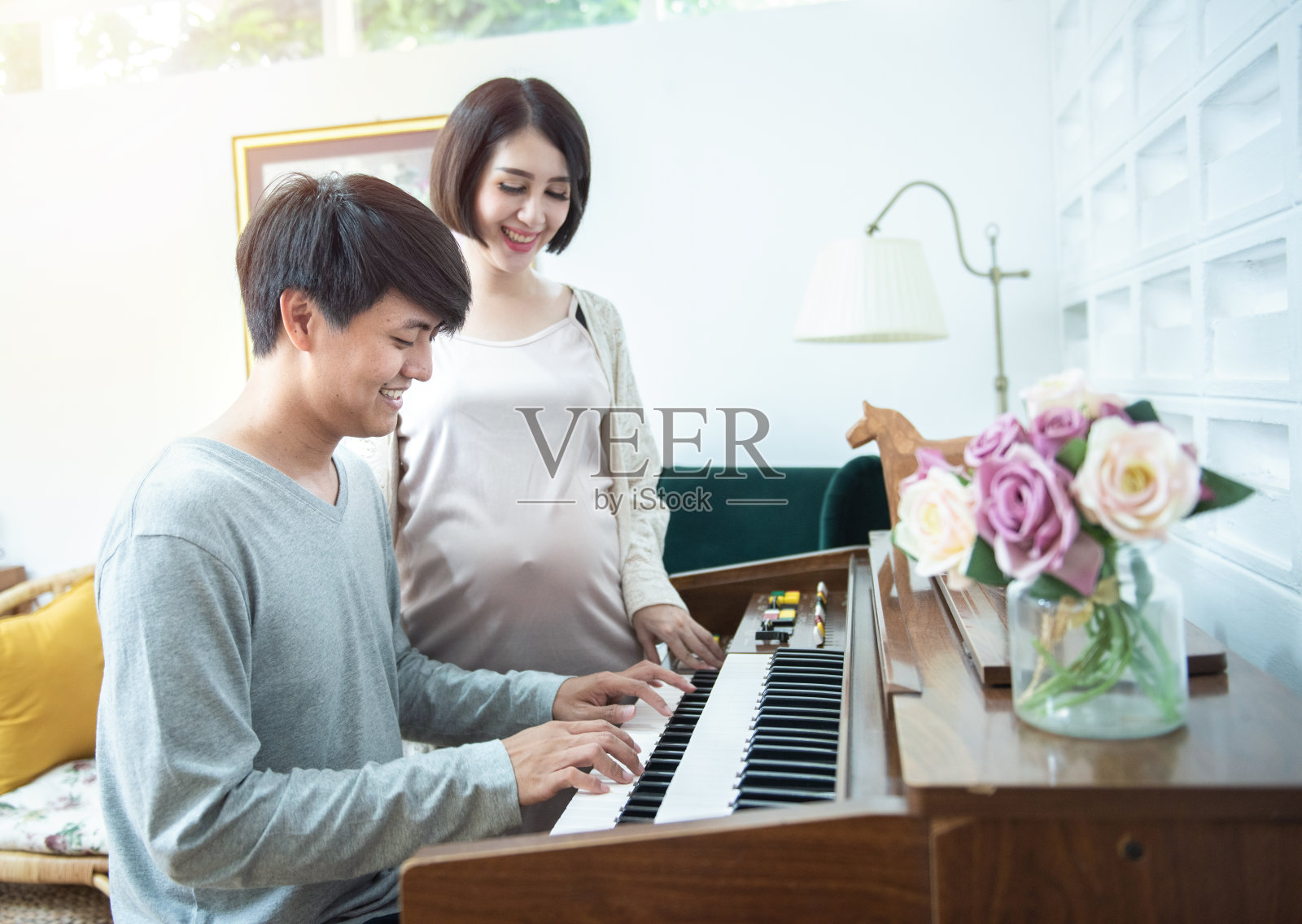 快乐年轻的亚洲孕妇站在男人的一边在家弹钢琴。照片摄影图片