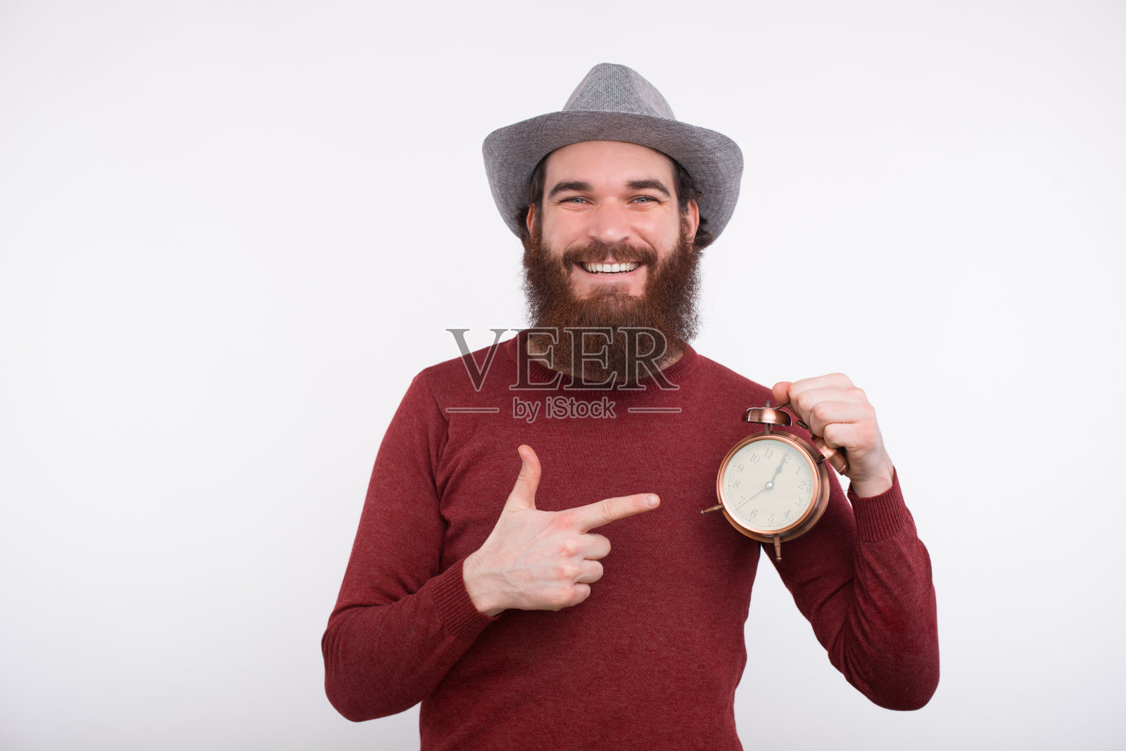 大胡子男子戴着一顶帽子对着镜头微笑，手里拿着一个指针指向镜头的时钟。照片摄影图片