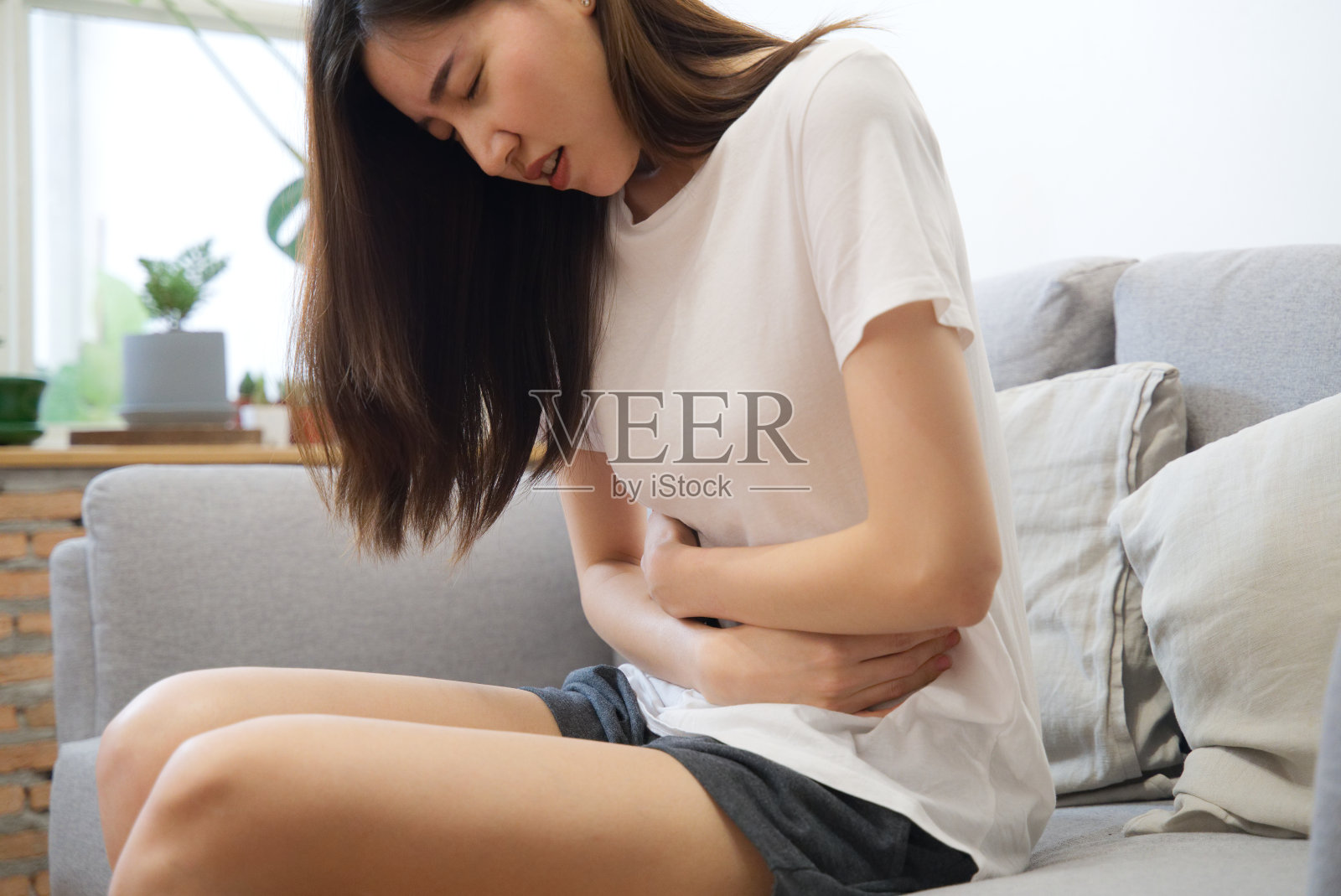 一个亚洲女孩正在经期，她坐在沙发上，感觉肚子很痛。它被称为女性疼痛或月经综合症。她抓住自己的腹部，在痉挛中扭动。照片摄影图片