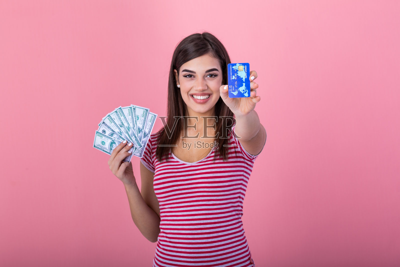照片上的快乐年轻女子站在孤立的粉红色背景。一边看一边拿着钱和信用卡。照片摄影图片