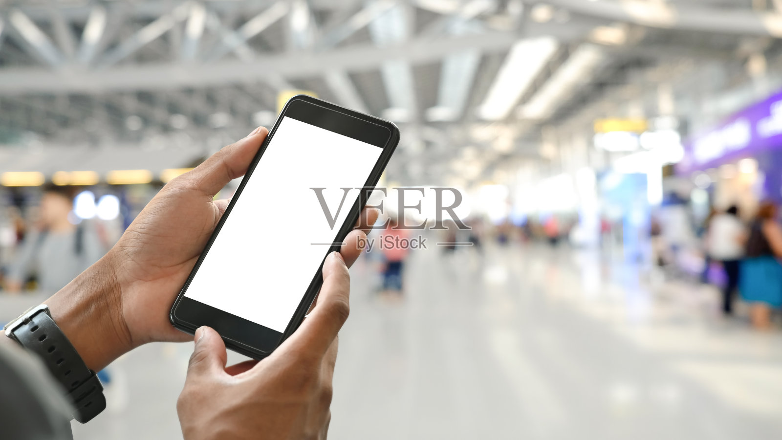 一名聪明男子的手拿着一部裁剪过的黑色智能手机，在拥挤的国际机场上空，黑屏是模糊的背景。照片摄影图片