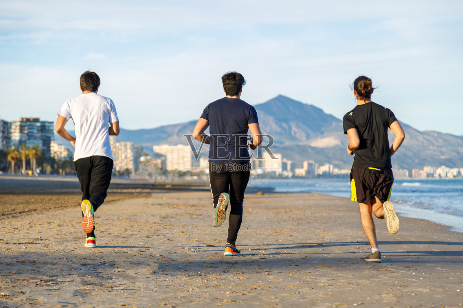 三个年轻人在日落时沿着海滩跑步的后视图照片摄影图片