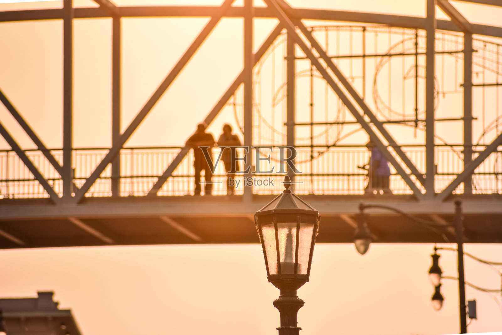 一对面目全非的夫妇凝视着夕阳下一座桥上的景色。前景是街灯。照片摄影图片