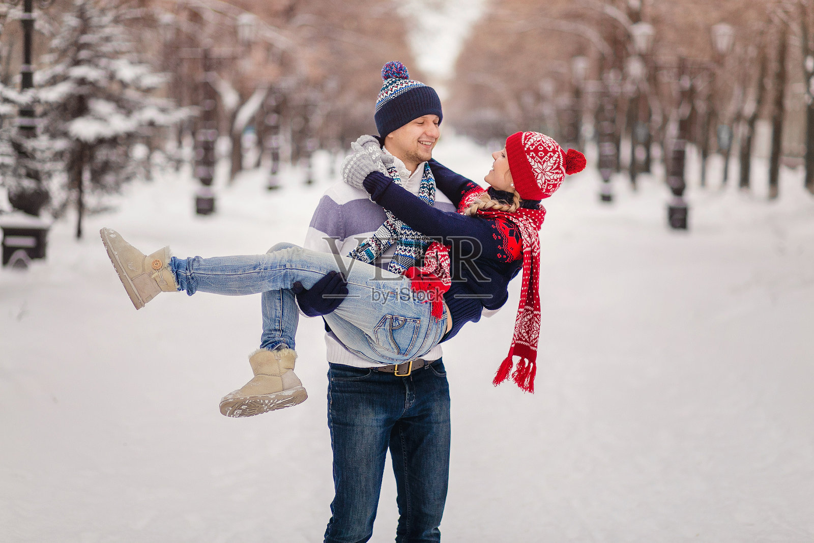 微笑的男人站起来，她的女人戴着帽子和围巾拥抱在下雪的冬天公园照片摄影图片