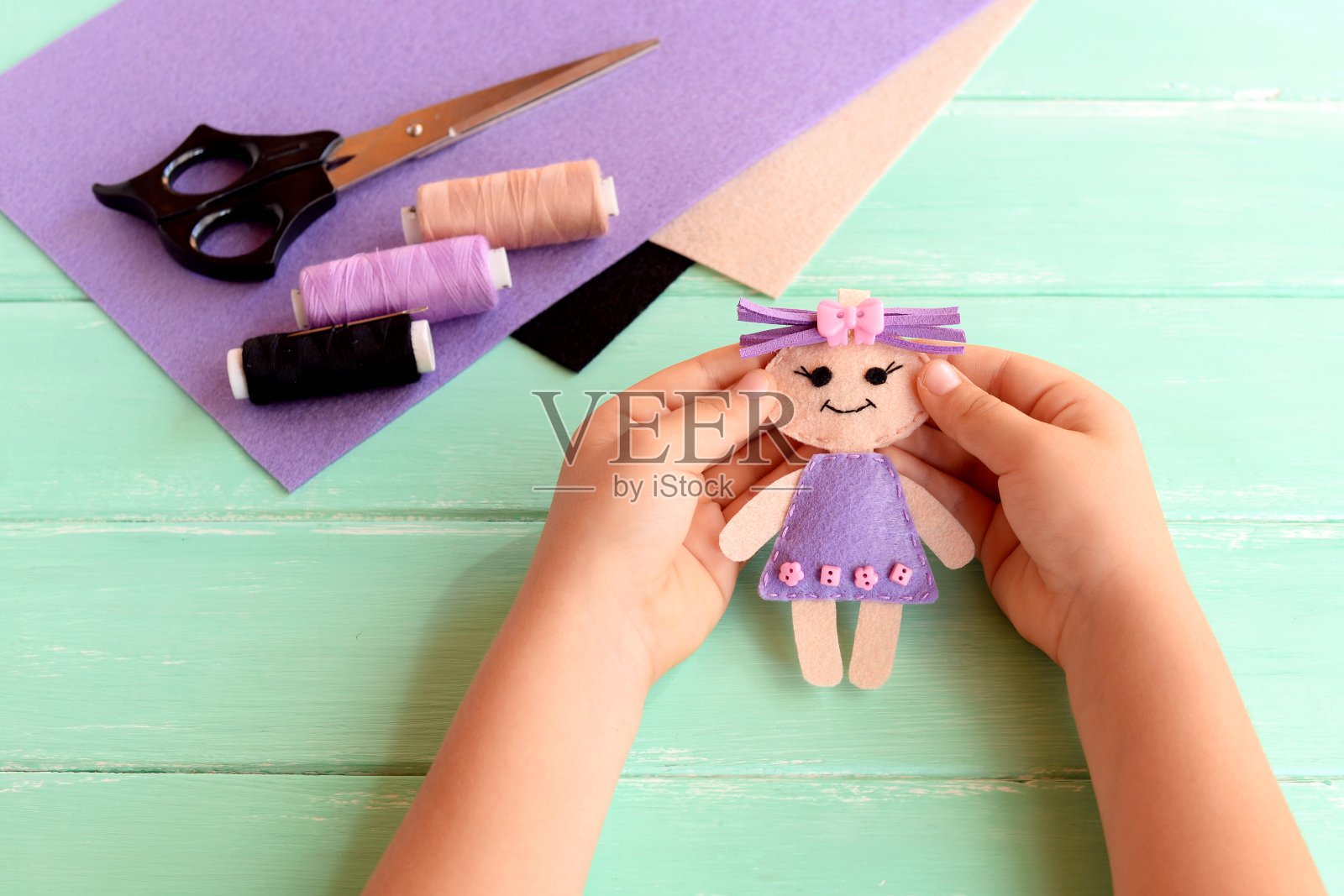 一个小孩手里拿着一个小娃娃。娃娃是用毛毡缝制的。把剪刀、线和毛毡放在一张木桌上。可爱的织物艺术和手工艺的想法，为孩子照片摄影图片