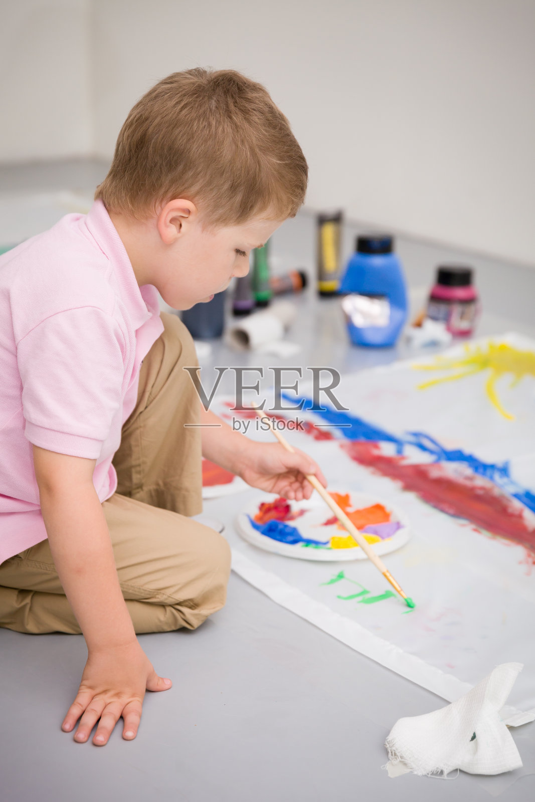 可爱的小男孩坐在地板上，用五颜六色的颜料画画。孩子在做旗子工艺品。教育，创造力，学校活动。照片摄影图片