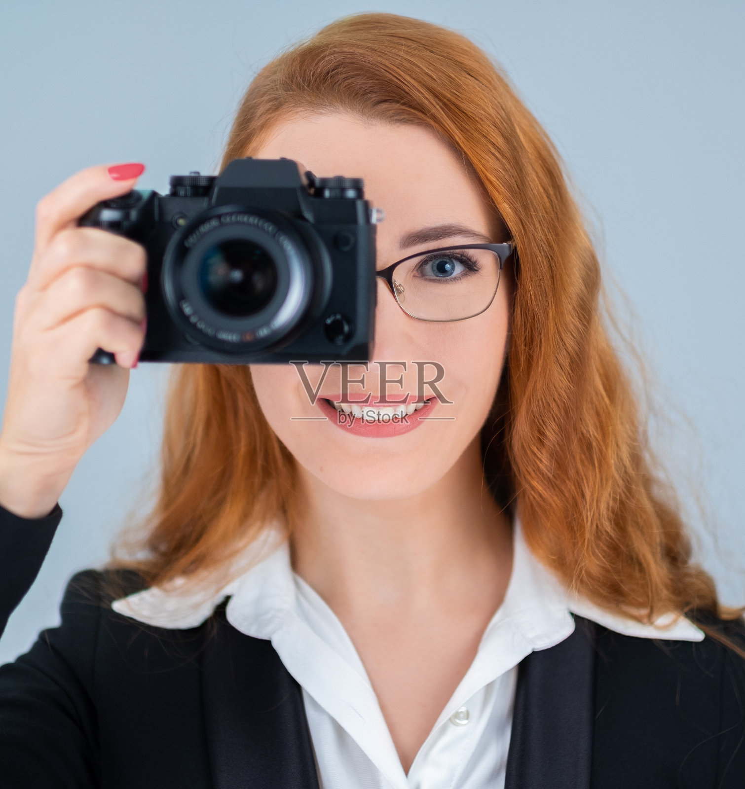 年轻的红发女人拿着照相机。她戴着眼镜，穿着西装。照片摄影图片