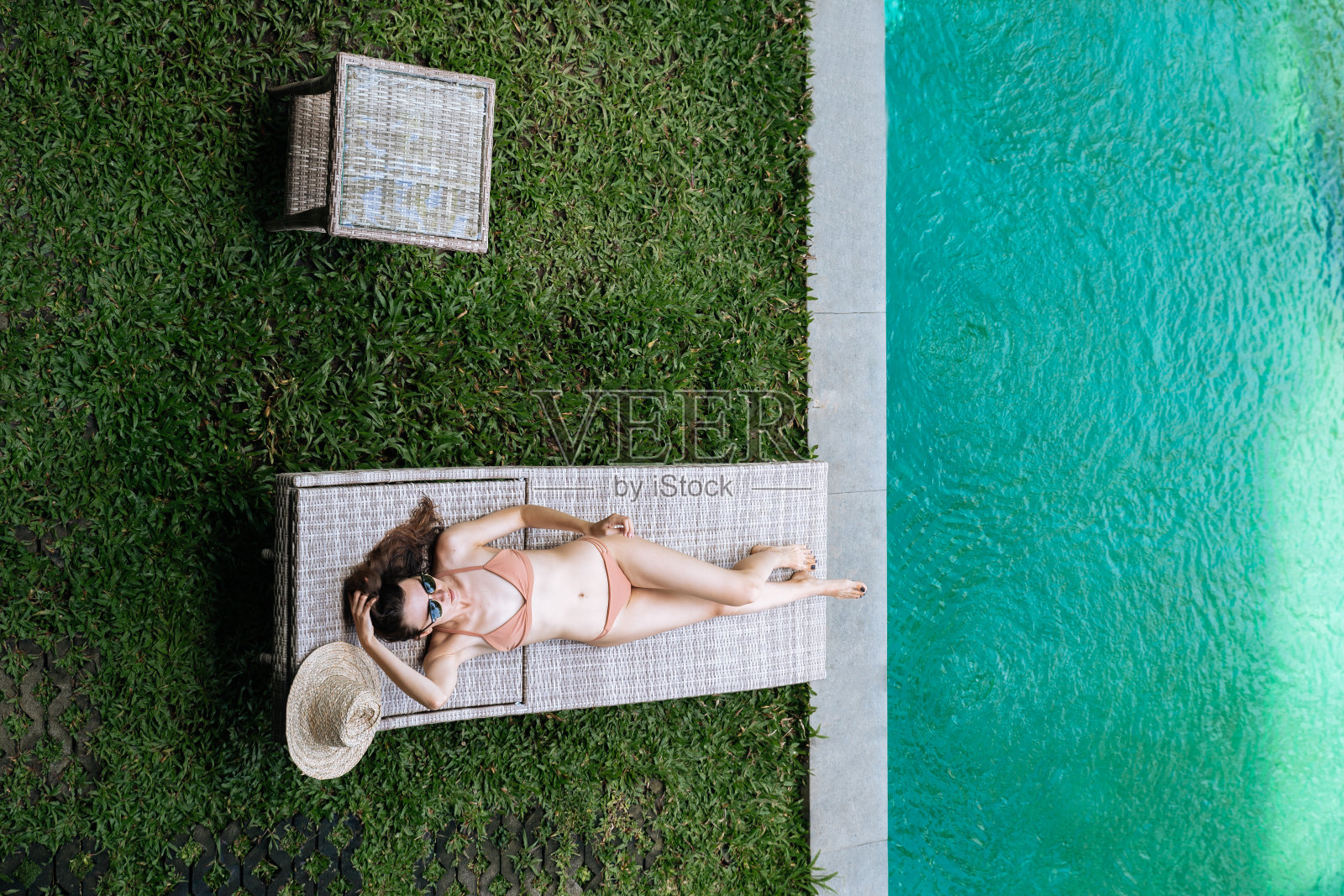 俯视图苗条的女人在米色比基尼采取日光浴在豪华泳池附近的热带度假躺椅上。照片摄影图片