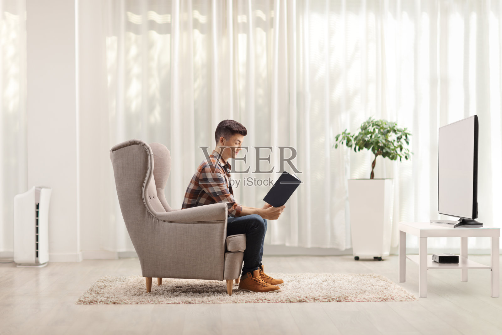 一个年轻人坐在扶手椅上，在电视机前看书的侧面照片照片摄影图片