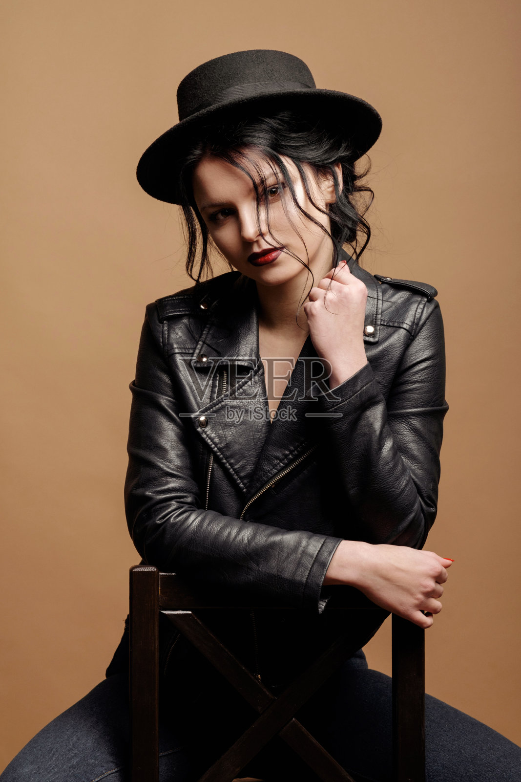 年轻美女时装模特在米色背景上戴着肖像帽子，穿着黑色皮夹克，坐在椅子上照片摄影图片