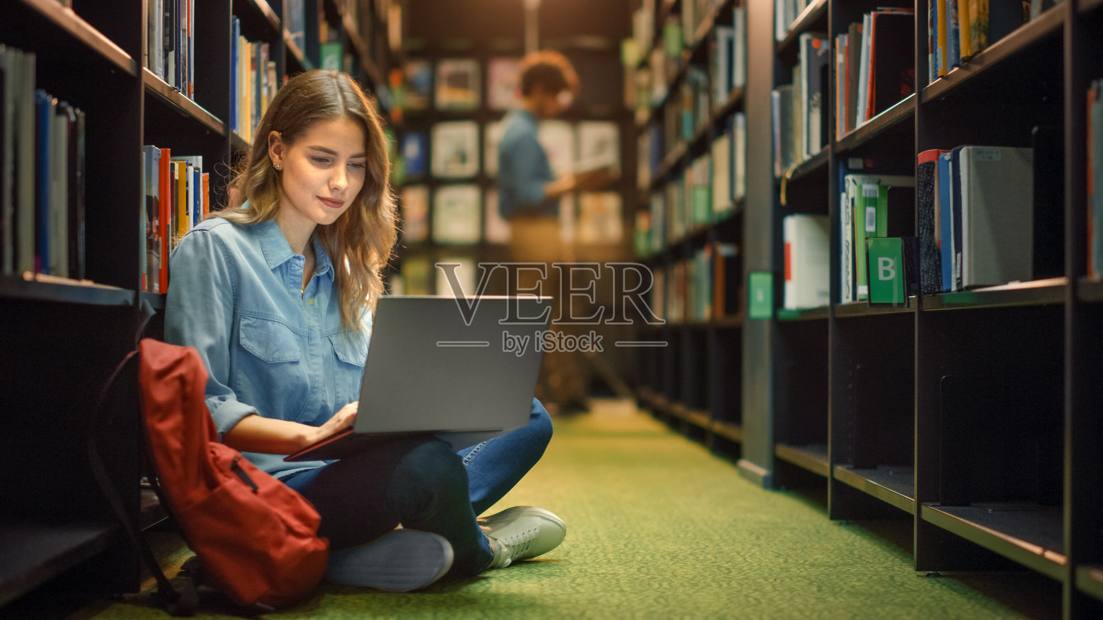 大学图书馆:天才美丽的白人女孩坐在地板上，使用笔记本电脑，为论文写笔记，论文，为课堂作业学习。不同的学生群在学习，为考试而学习。照片摄影图片