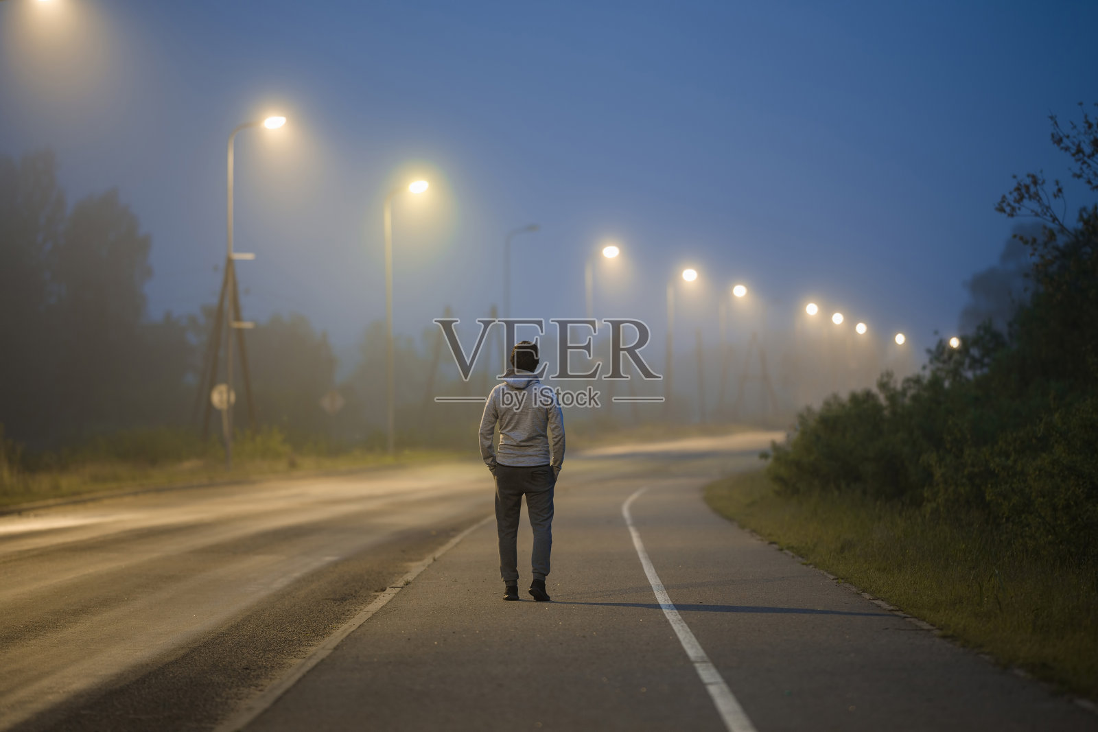 一个年轻人在夏夜的路灯下慢慢地走在长长的人行道上。花时间独自呆着。后视图。照片摄影图片