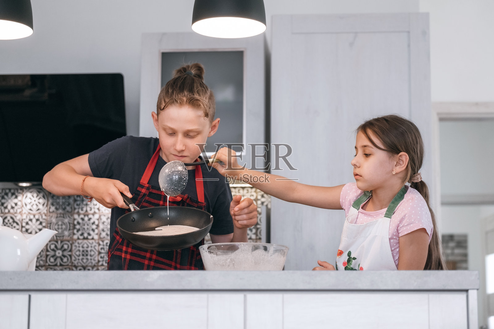 哥哥和妹妹穿着围裙，在家里的厨房里自制煎饼。女孩在热锅上揉面团。儿童家庭烹饪概念形象。照片摄影图片
