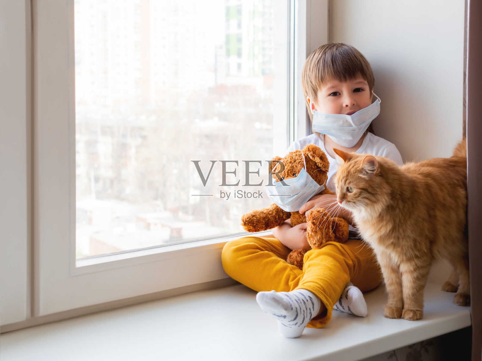 蹒跚学步的小男孩和泰迪熊坐在一起，都戴着医用口罩。孩子和可爱的姜黄色的猫。新型冠状病毒感染的宠物和儿童接受家庭隔离。照片摄影图片