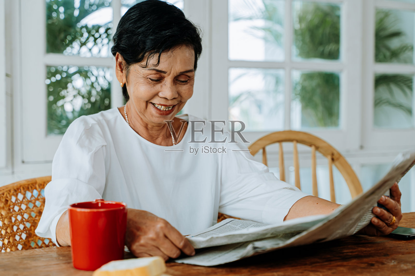 快乐的资深亚洲女性在休闲衣服阅读有趣的文章在报纸上，而在家里吃早餐。享受退休生活的老年妇女照片摄影图片