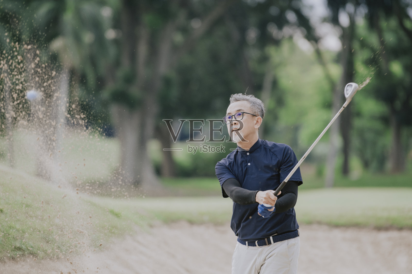 一位亚洲的中国资深高尔夫球手用他的铁棒在沙坑里打他的高尔夫球照片摄影图片