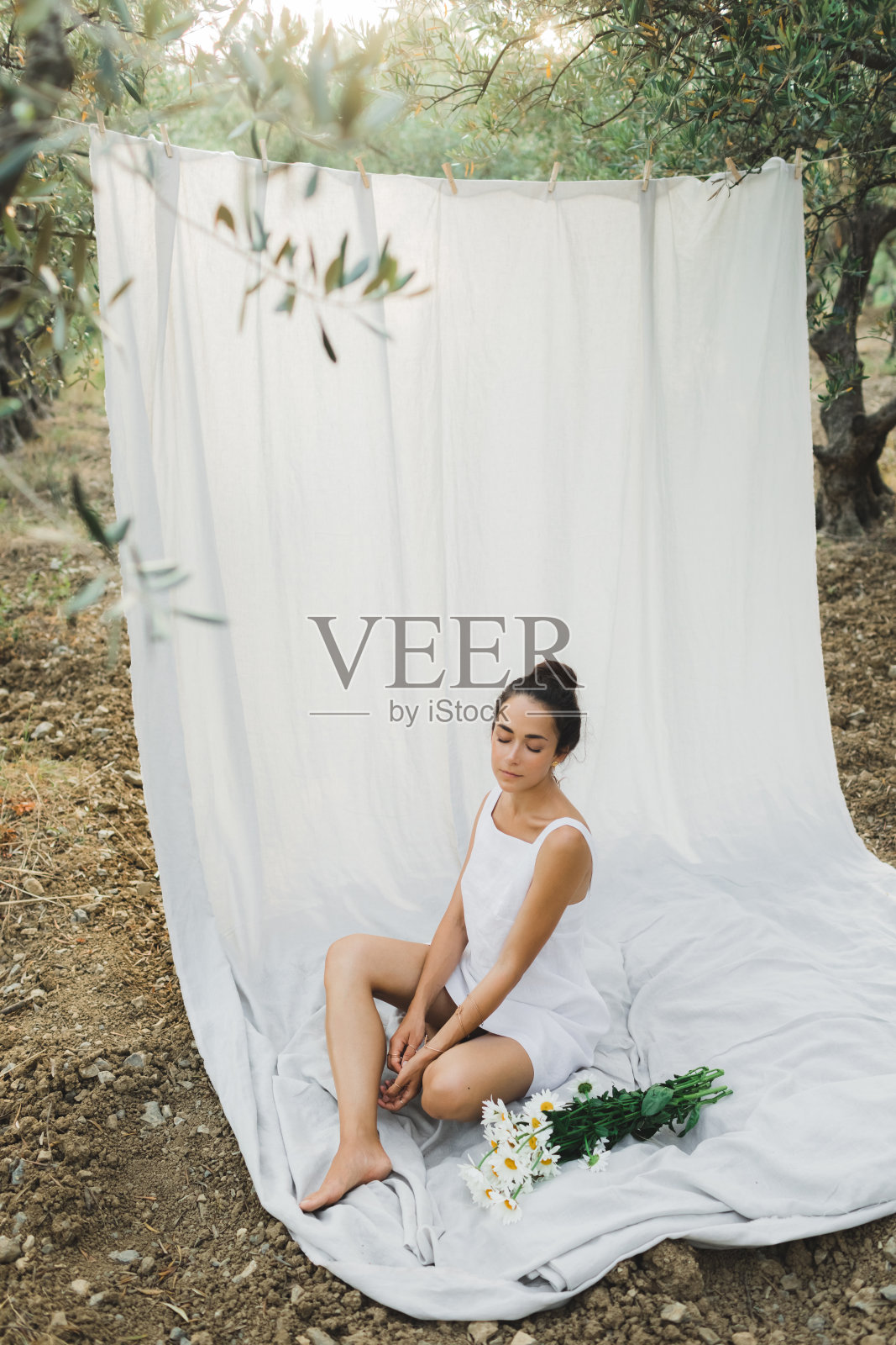 年轻的黑发女子的肖像在白色的夏季亚麻连衣裙的背景上悬挂纺织品布。放松休闲，橄榄花园野餐，一束洋甘菊。照片摄影图片