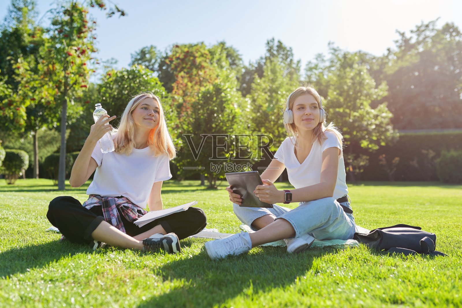 女生们坐在绿色的草坪上拿着数码平板电脑，喝着瓶装水照片摄影图片